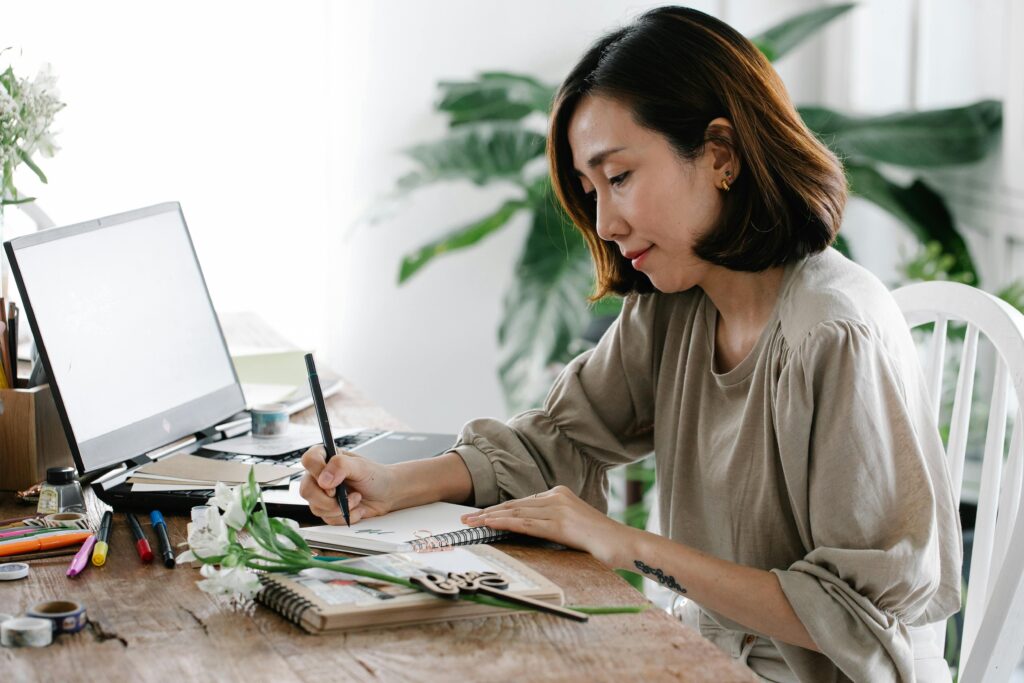 An adult woman writes in a notebook at a wooden desk with a laptop and flowers, creating a serene indoor scene.
